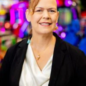 A woman in a suit stands confidently in front of a colorful carnival, with rides and decorations in the background.