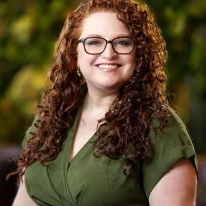 A woman with curly red hair and glasses smiles at the camera, showcasing her vibrant hair and stylish eyewear.