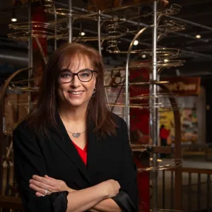 A woman with glasses and a red shirt stands in front of an abstract metal sculpture, showcasing her surroundings.