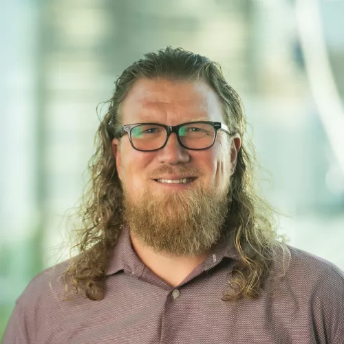 Man with shoulder length blonde hair wearing glasses and pink collared shirt