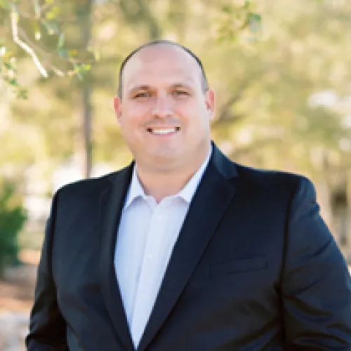  A man in a suit and tie stands confidently in front of lush green trees