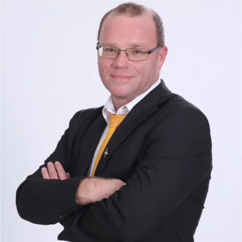 Armand Dore wearing a black suit with a yellow tie standing in front of a grey background, arms crossed