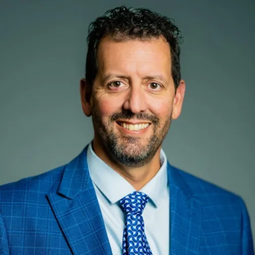 Jason smiling at the camera in front of a dark background, wearing a blue suit and tie