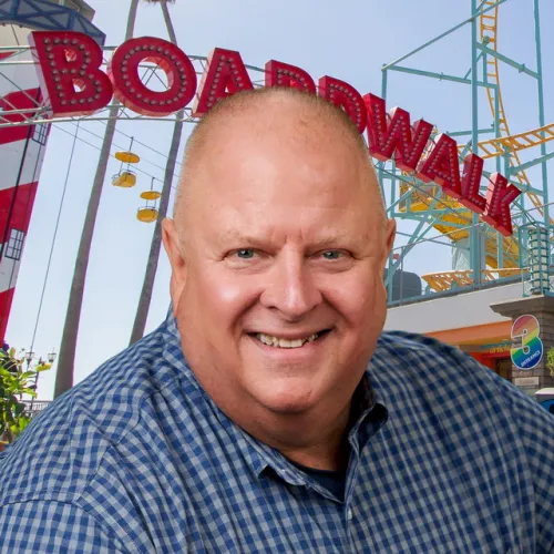 Robert smiling at the camera in front of the Santa Cruz Beach Boardwalk entrance archway, wearing a blue chequered shirt