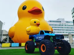 An inflatable duck sits in front of a monster truck at a sporting events stadium.
