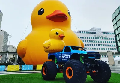 An inflatable duck sits in front of a monster truck at a sporting events stadium.