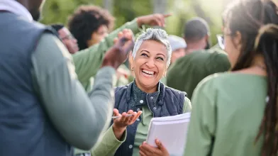 Woman with short white hair holding pen and paper, smiling with co-workers